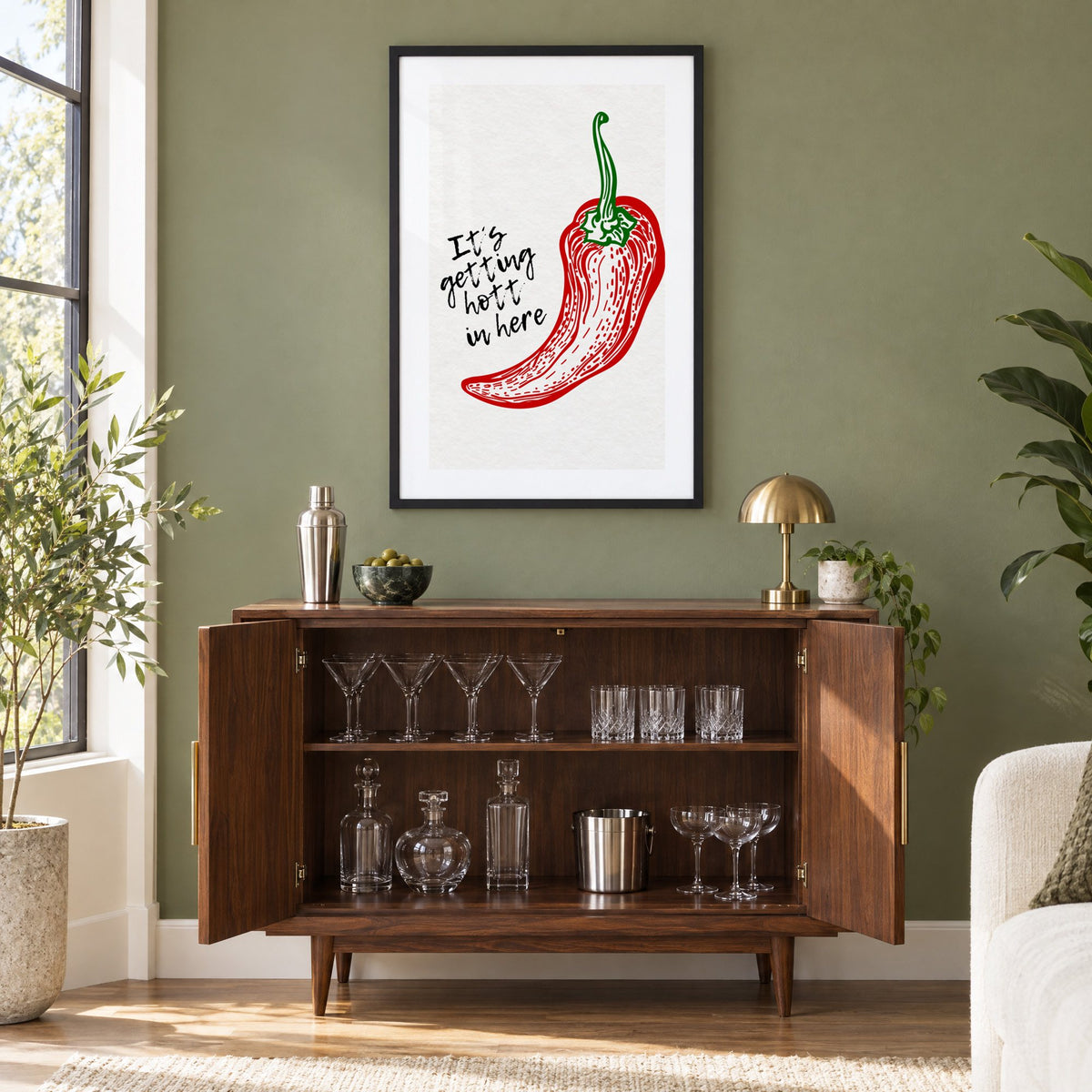 It's getting hott red chili engraving print in a black frame on an olive green wall above a walnut bar cabinet with glassware