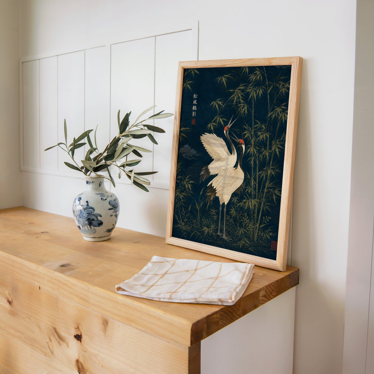 red-crowned cranes and bamboo on navy print in a pine frame on a wooden counter beside a blue and white porcelain vase