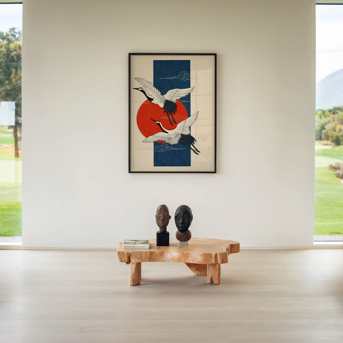 red-crowned cranes on navy with red sun print in a black frame above a live-edge wood coffee table between garden windows