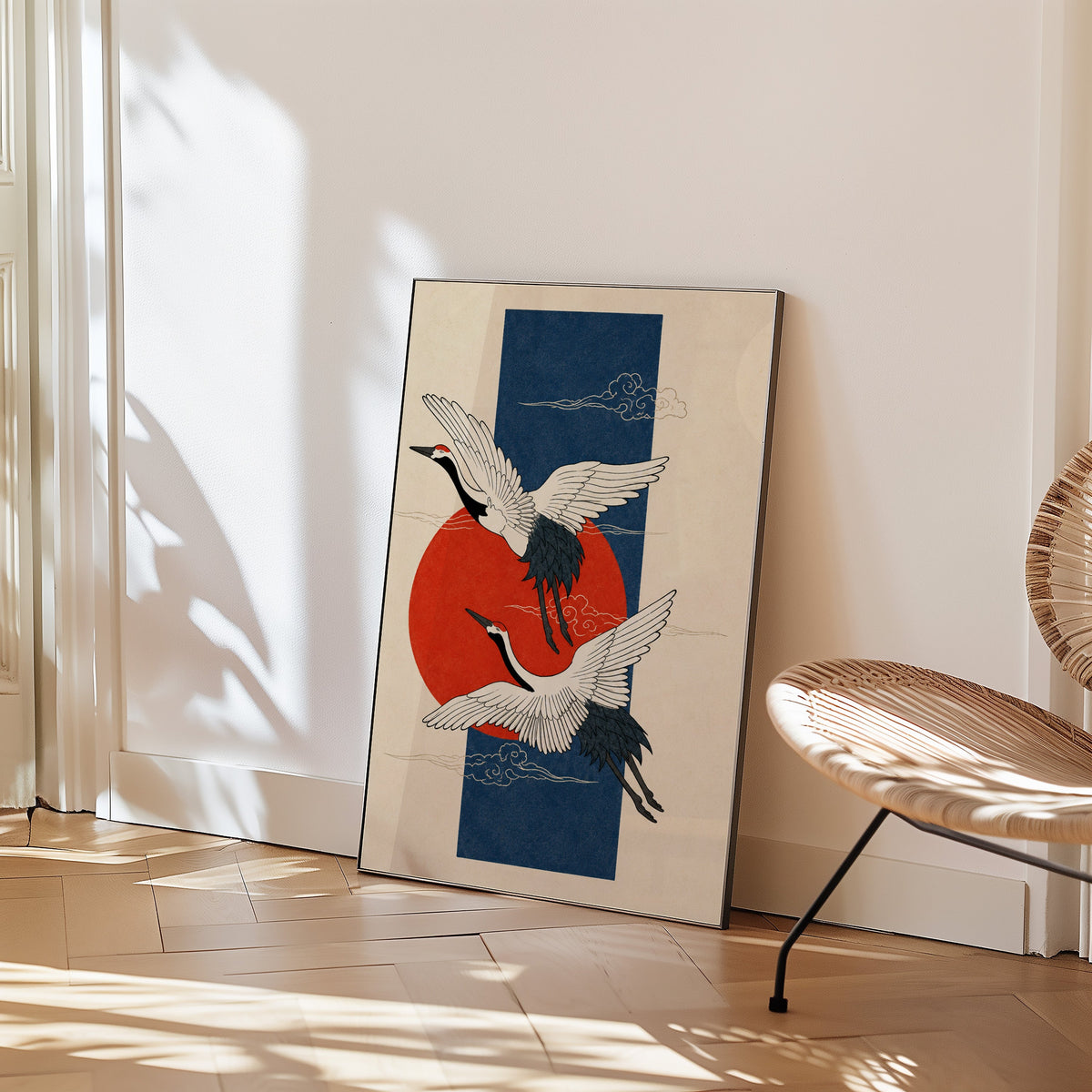 red-crowned cranes on navy with red sun print in a black frame leaning on a herringbone floor beside a woven rattan chair