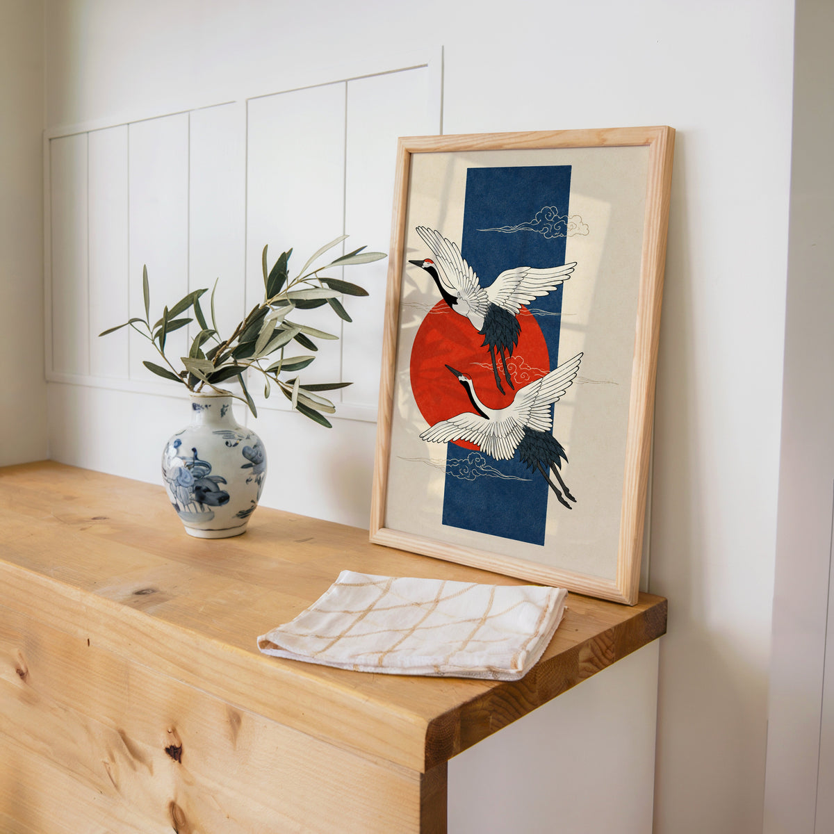 red-crowned cranes on navy with red sun print in a pine frame on a wooden counter beside a blue and white porcelain vase