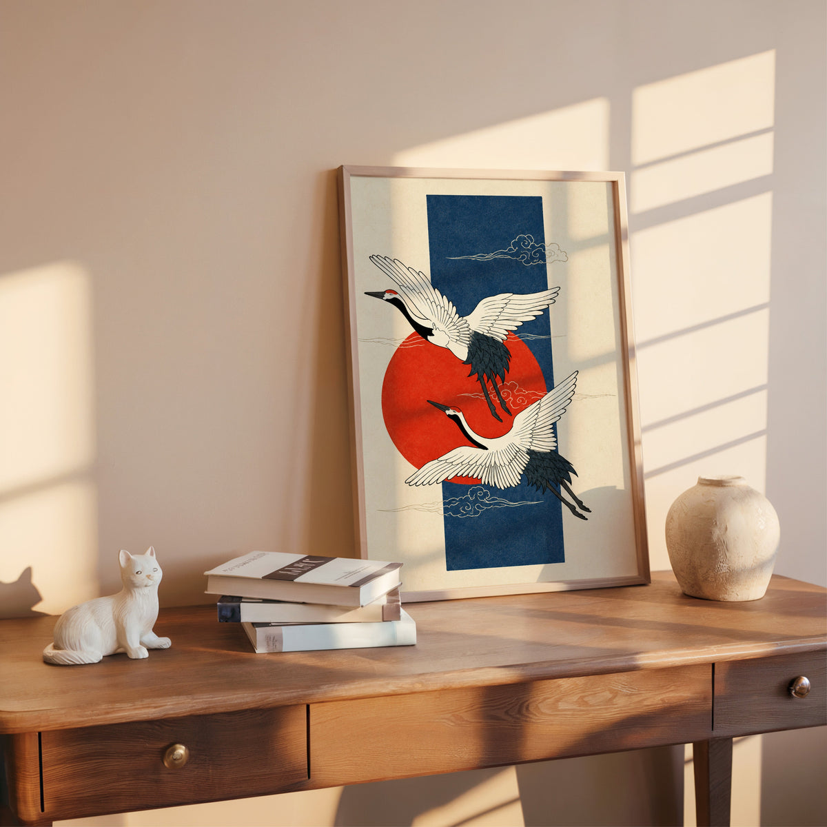red-crowned cranes on navy with red sun print shown in a rose gold frame leaning on a wooden writing desk with books