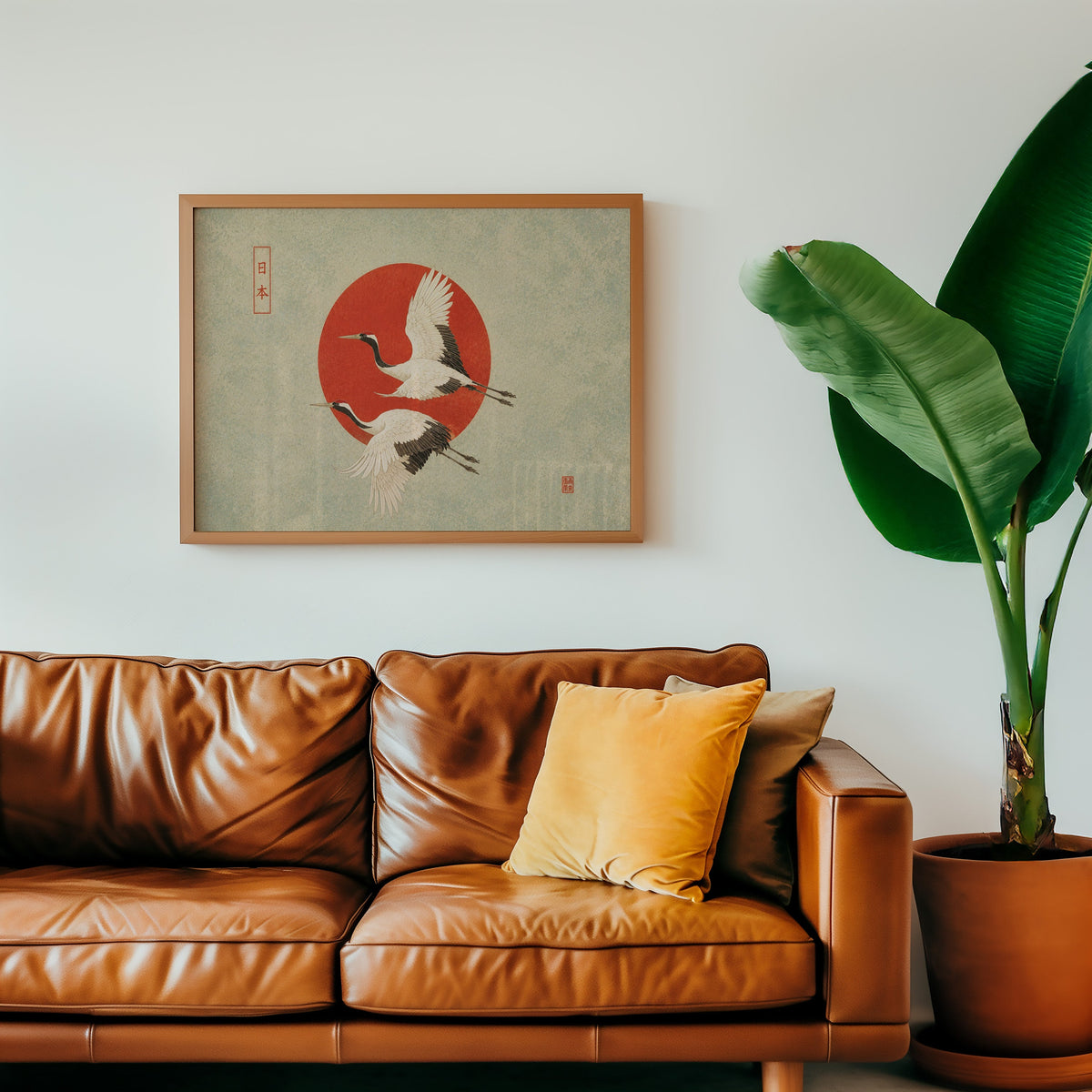 Red-crowned cranes flying past a red sun print in a warm oak frame above a cognac leather sofa with a mustard pillow