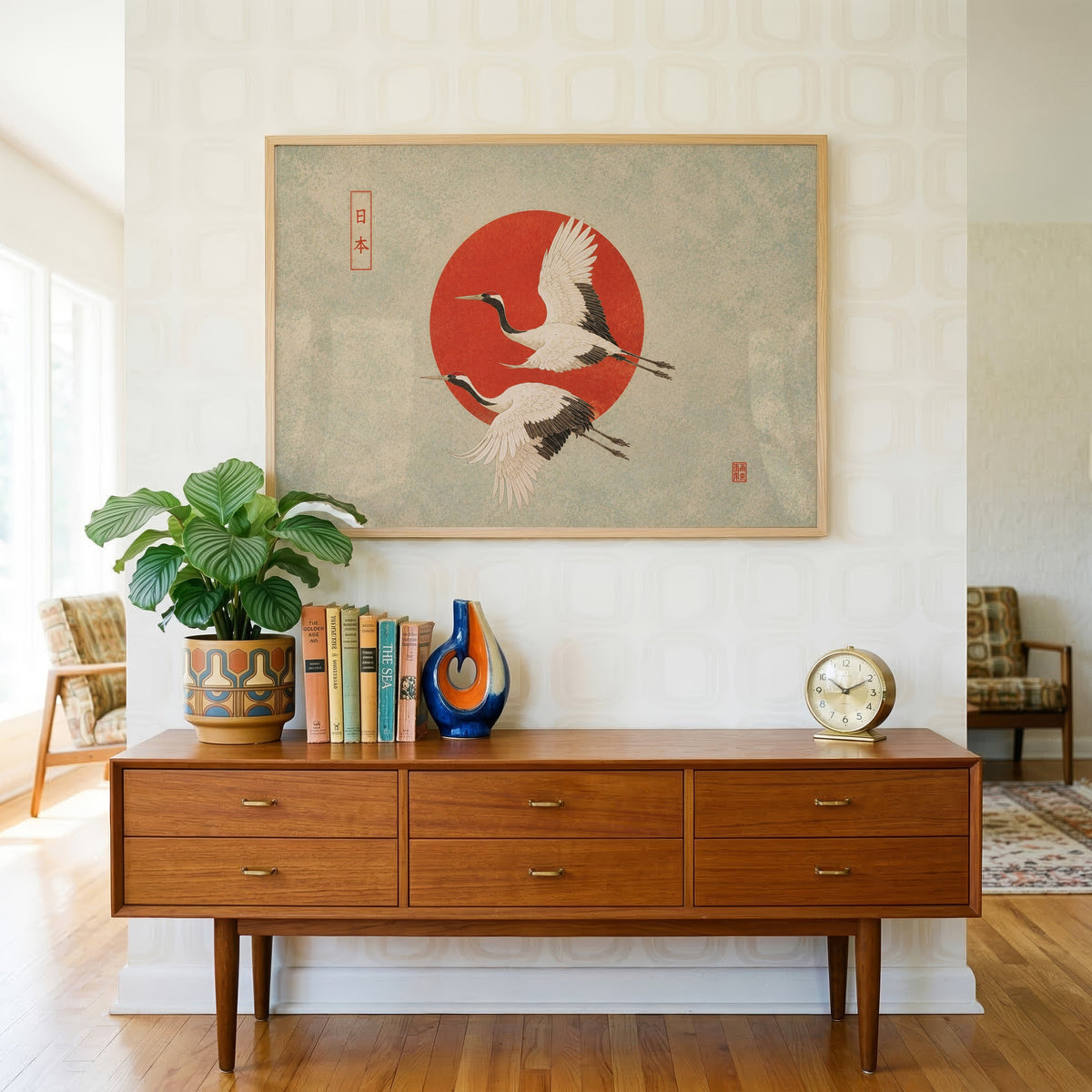 Red-crowned cranes flying past a red sun print in a light oak frame above a teak credenza with books and a brass clock