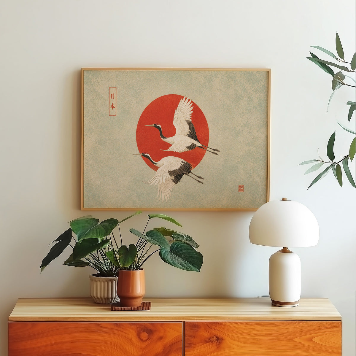Red-crowned cranes flying past a red sun print in a light oak frame above a wood sideboard with a white dome lamp