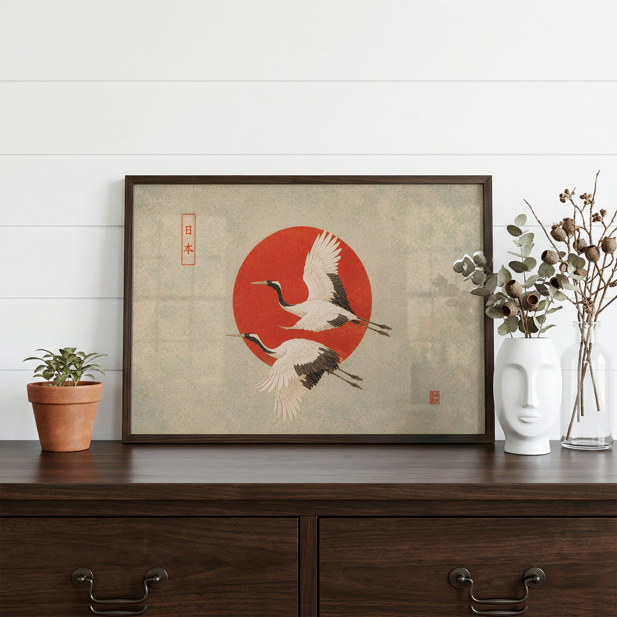 Red-crowned cranes flying past a red sun print in a dark walnut frame on a dark wood dresser beside a white face vase