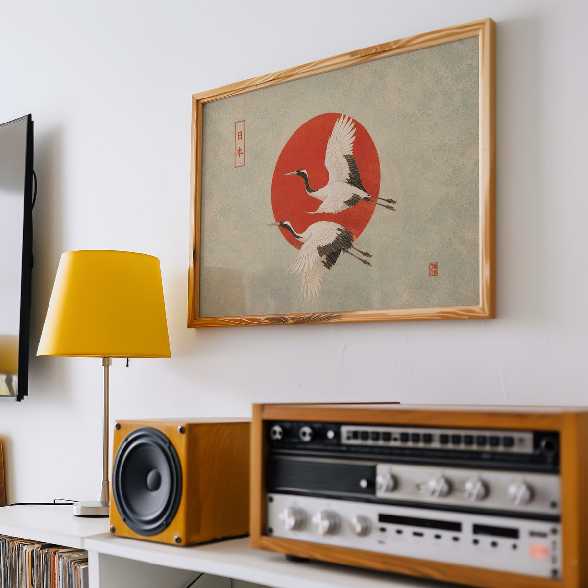 Red-crowned cranes flying past a red sun print in a pine frame above a vintage stereo and a yellow table lamp