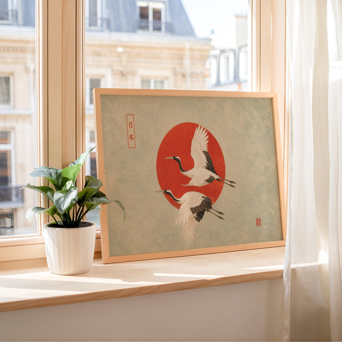 Red-crowned cranes flying past a red sun print in a light wood frame leaning on a sunny windowsill by sheer curtains
