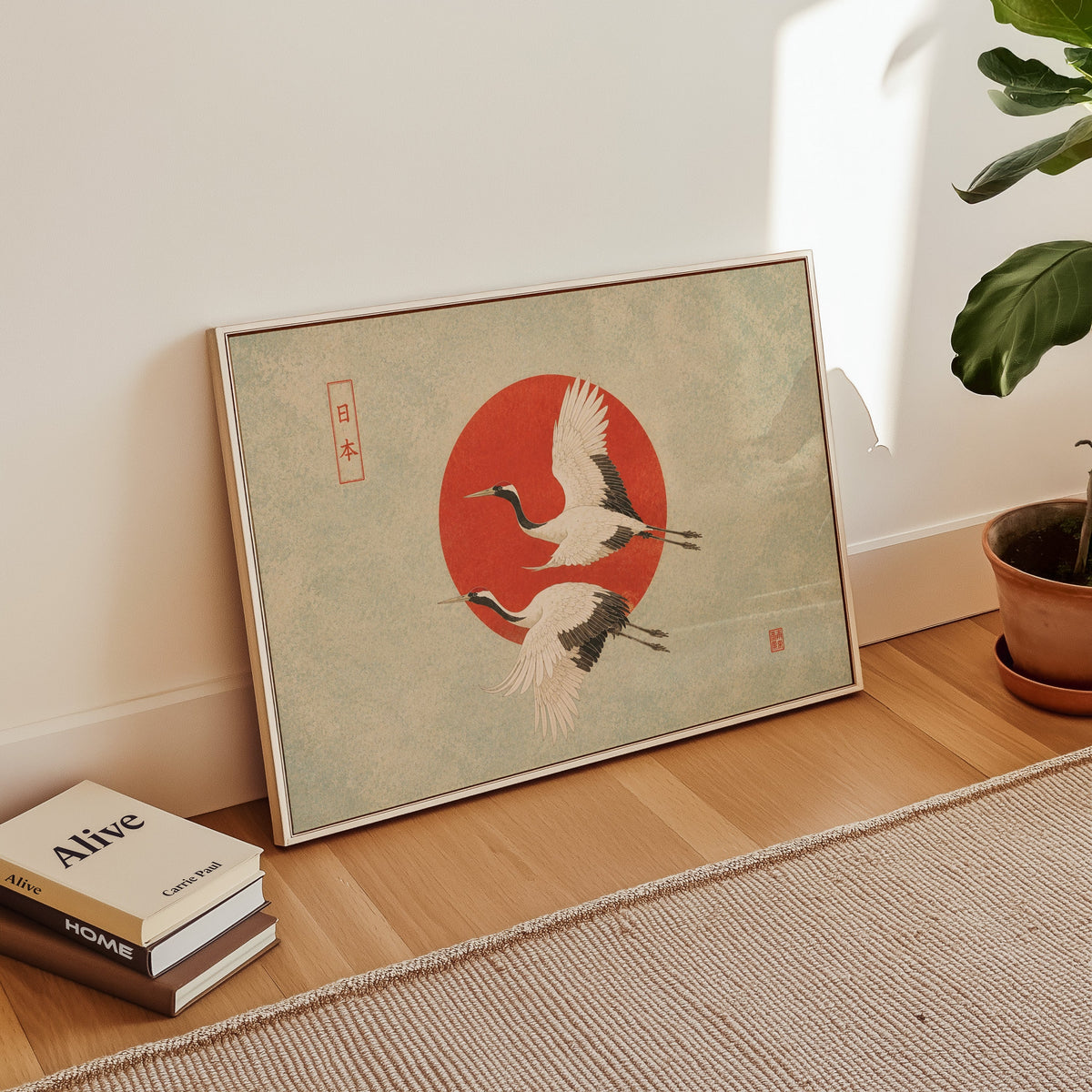 Red-crowned cranes flying past a red sun print in a white floater frame leaning on a wood floor beside stacked books