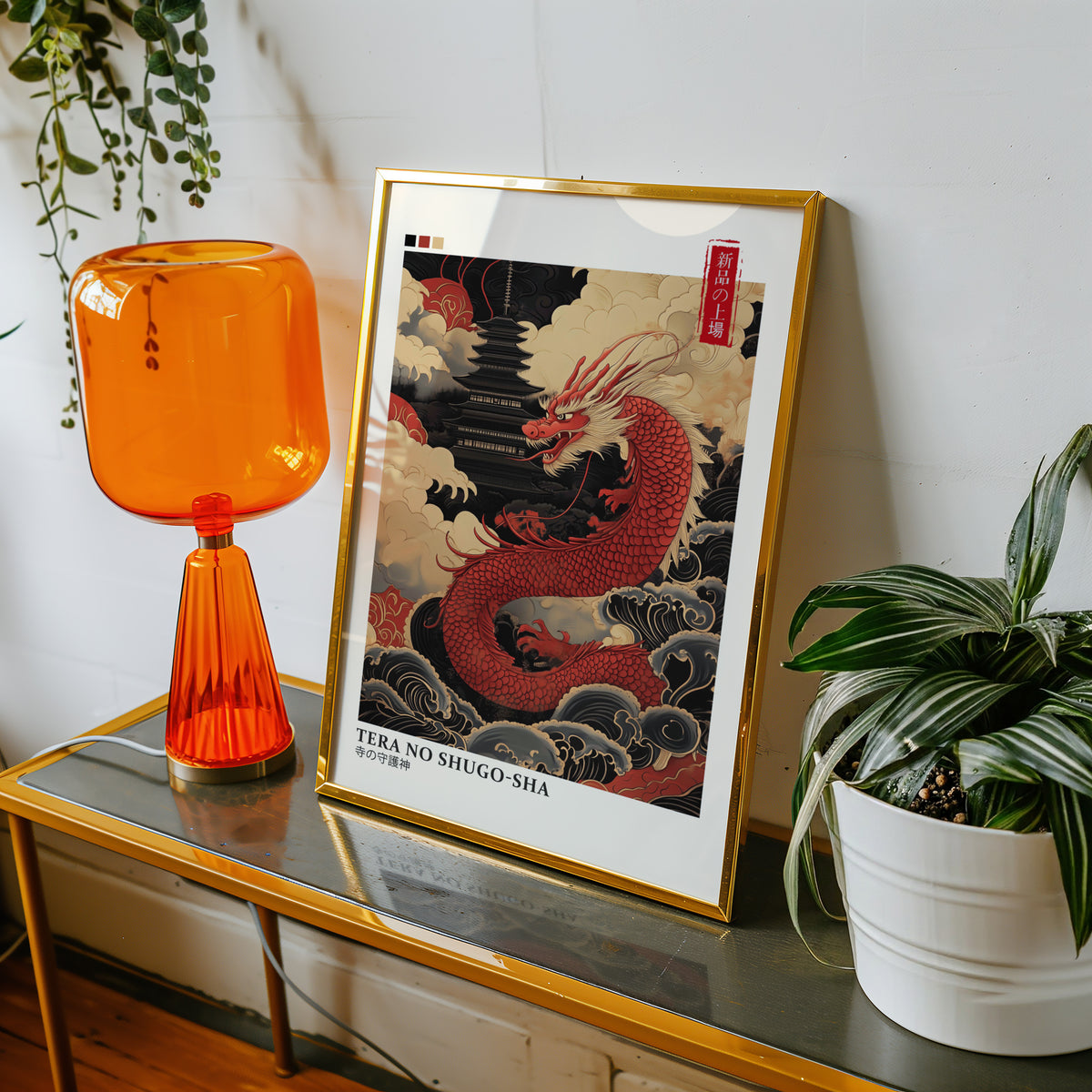 Red dragon guarding black pagoda print shown in a gold frame on a console beside an orange glass lamp