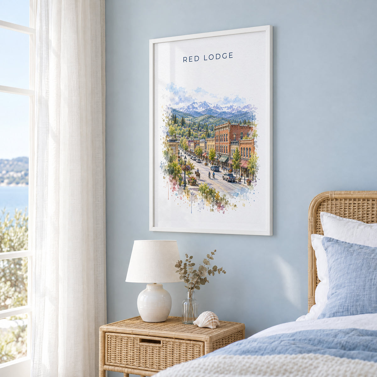 Red Lodge, Montana watercolor print in a white frame on a pale blue wall beside a rattan bed in a coastal bedroom
