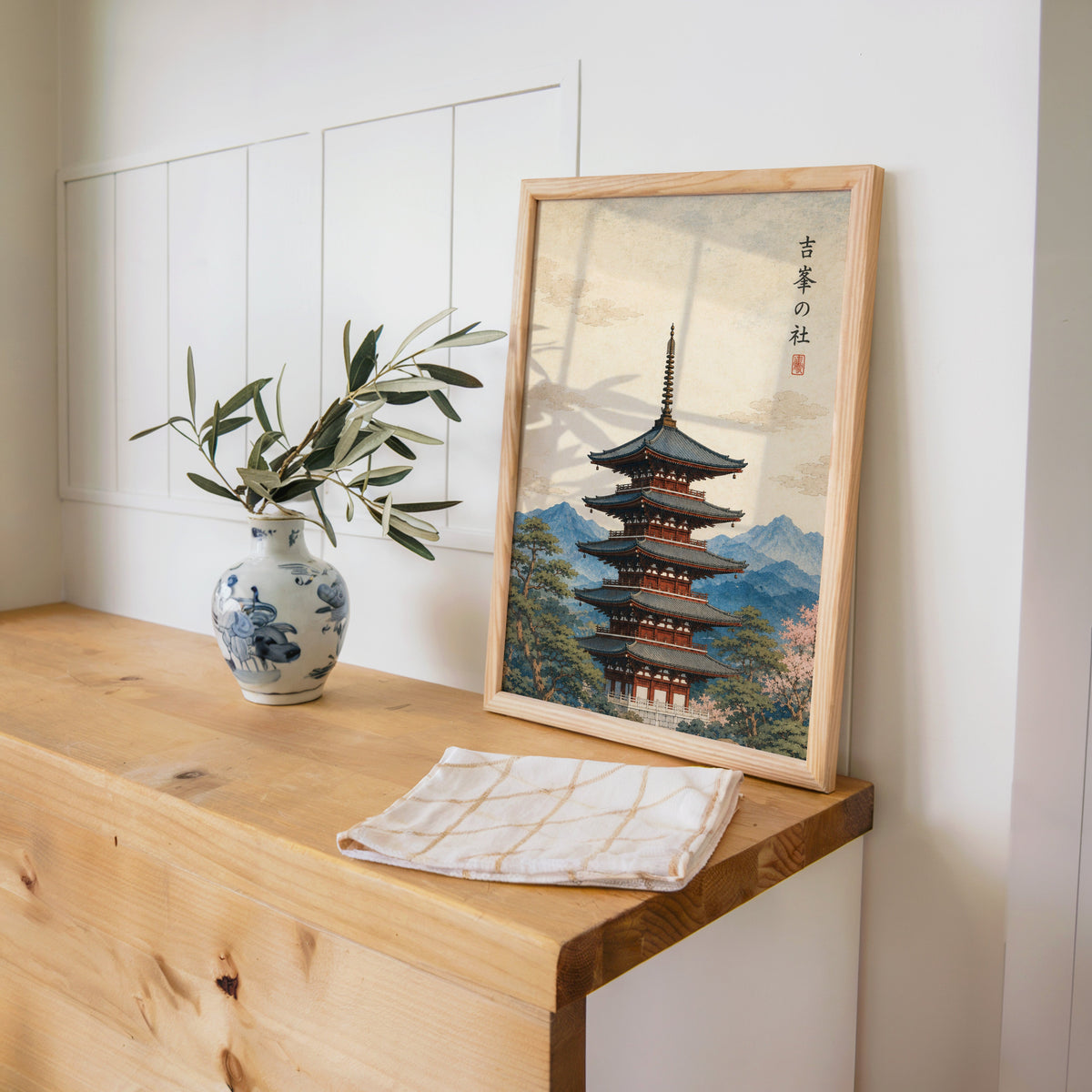 red pagoda with blue mountains print in a pine frame on a wooden counter beside a blue and white porcelain vase