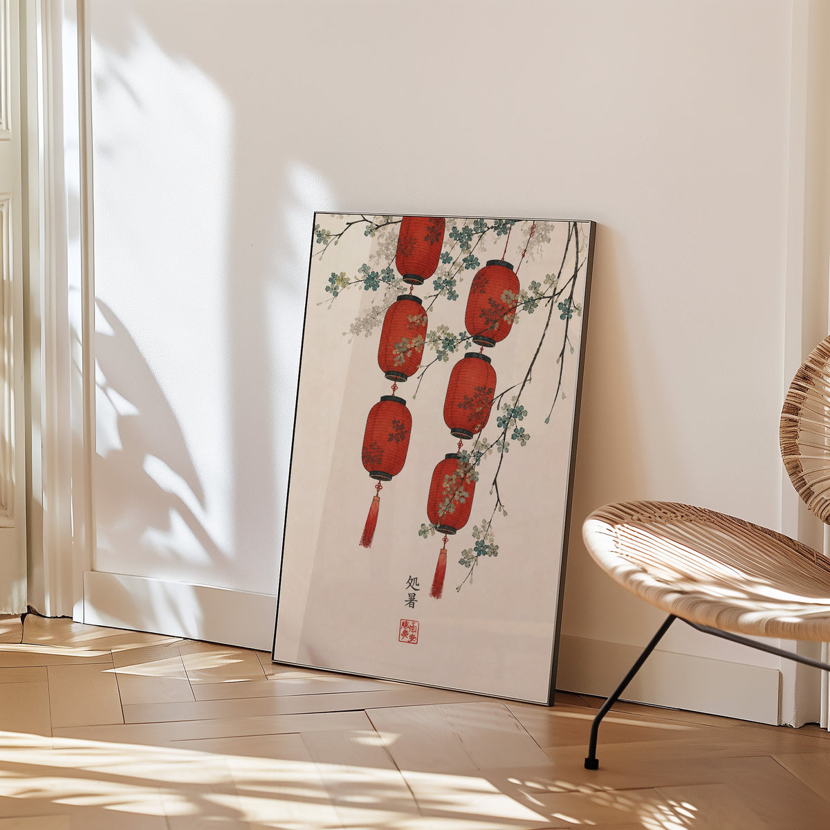 red paper lanterns with blossom branch print in a black frame leaning on a herringbone floor beside a woven rattan chair