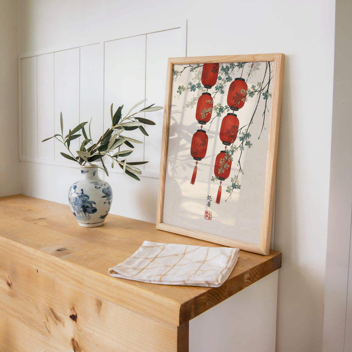 red paper lanterns with blossom branch print in a pine frame on a wooden counter beside a blue and white porcelain vase