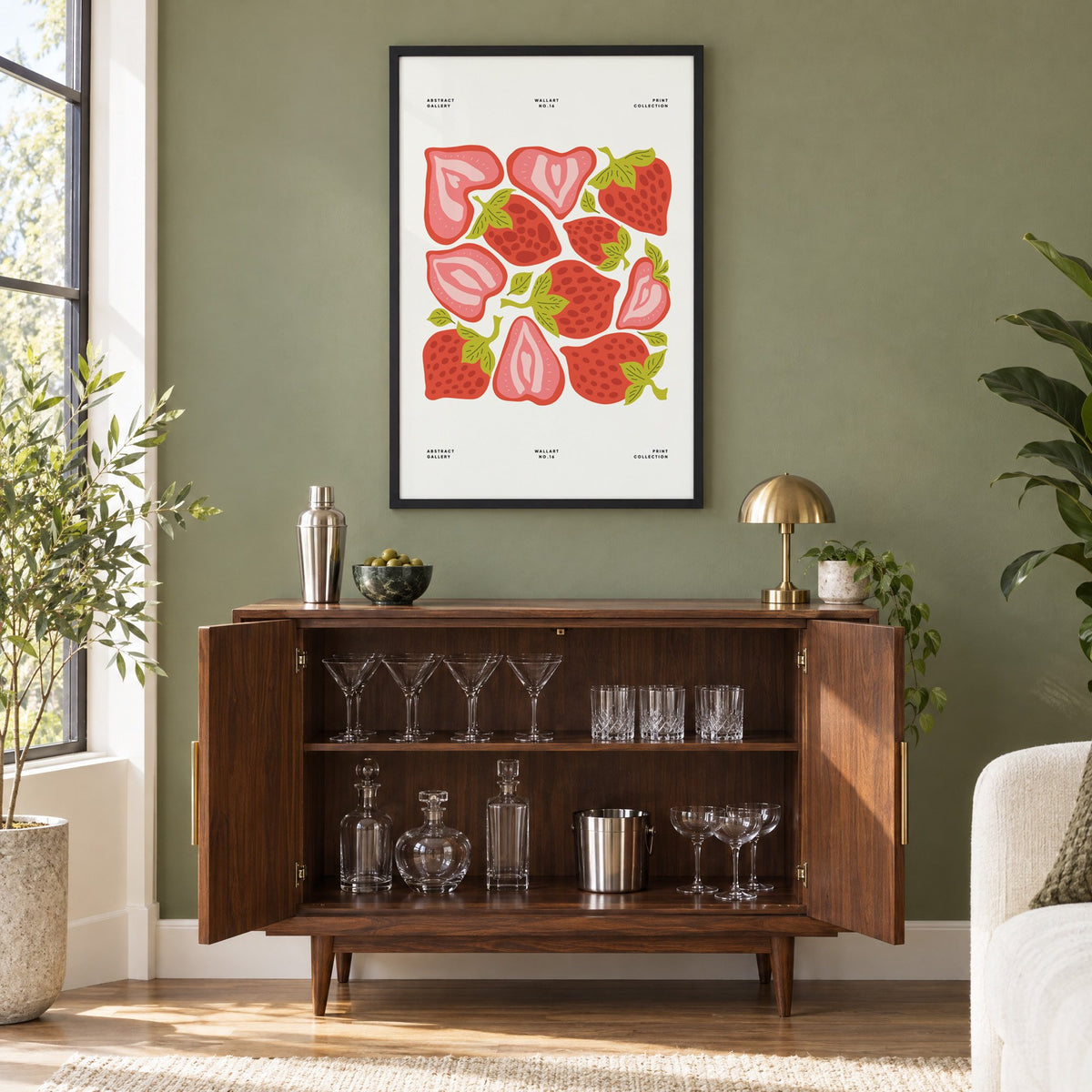 Red and pink strawberry halves pattern print in a black frame on an olive green wall above a walnut bar cabinet with glassware