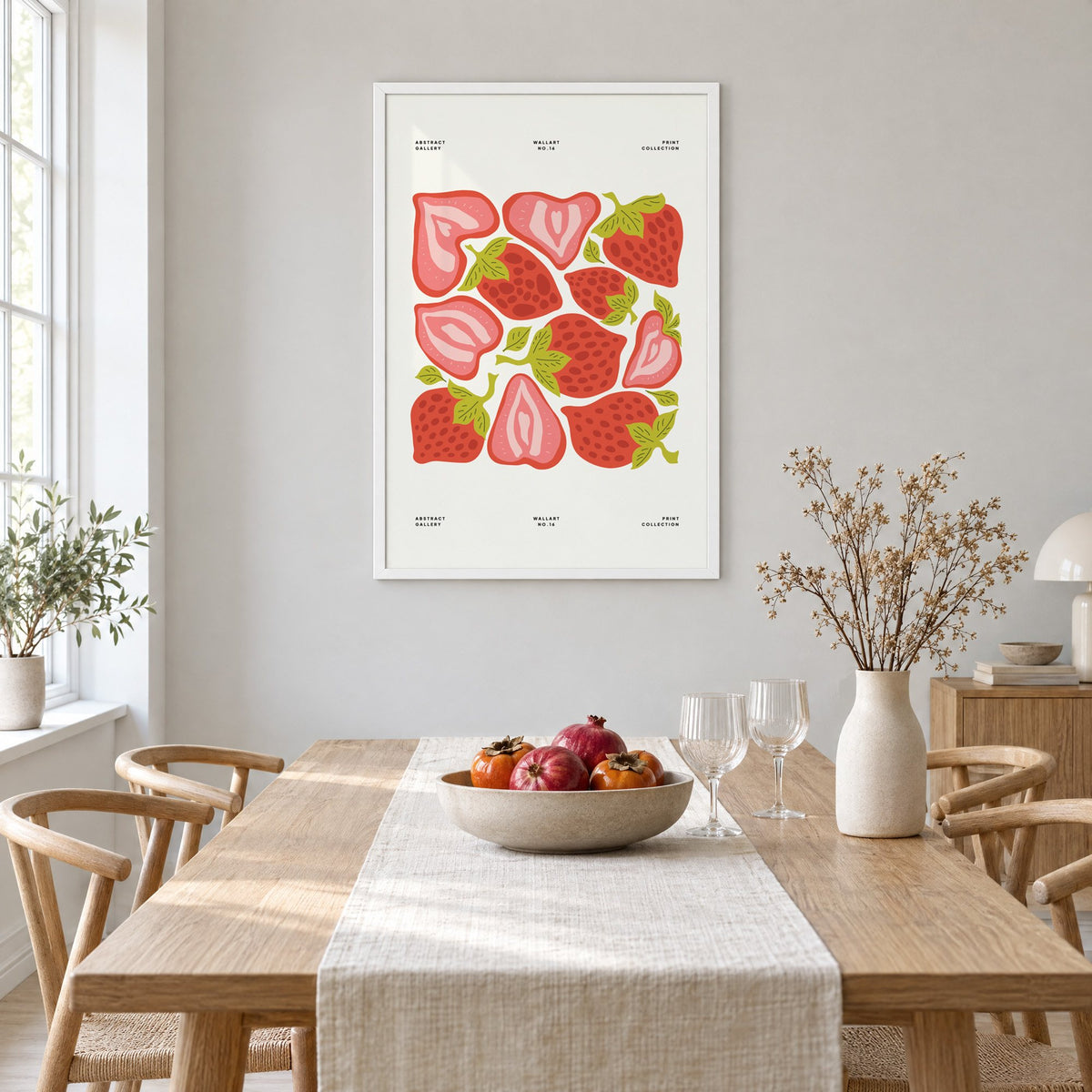 Red and pink strawberry halves pattern print in a white frame above a light oak dining table with a linen runner and fruit bowl