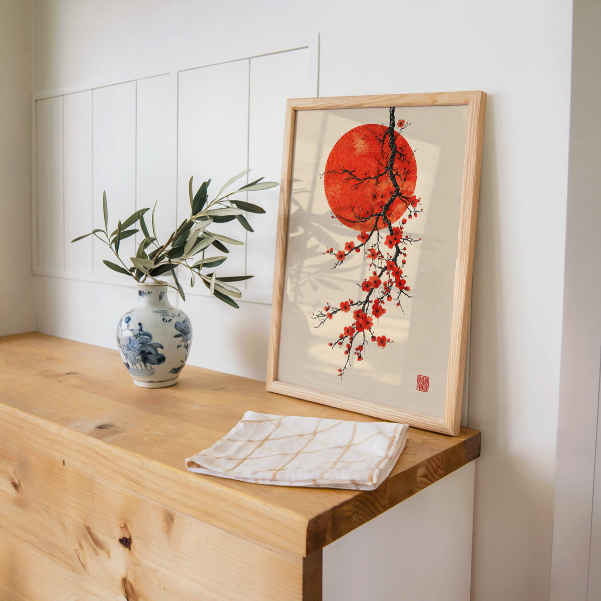 red blossom ink branch with red sun print in a pine frame on a wooden counter beside a blue and white porcelain vase