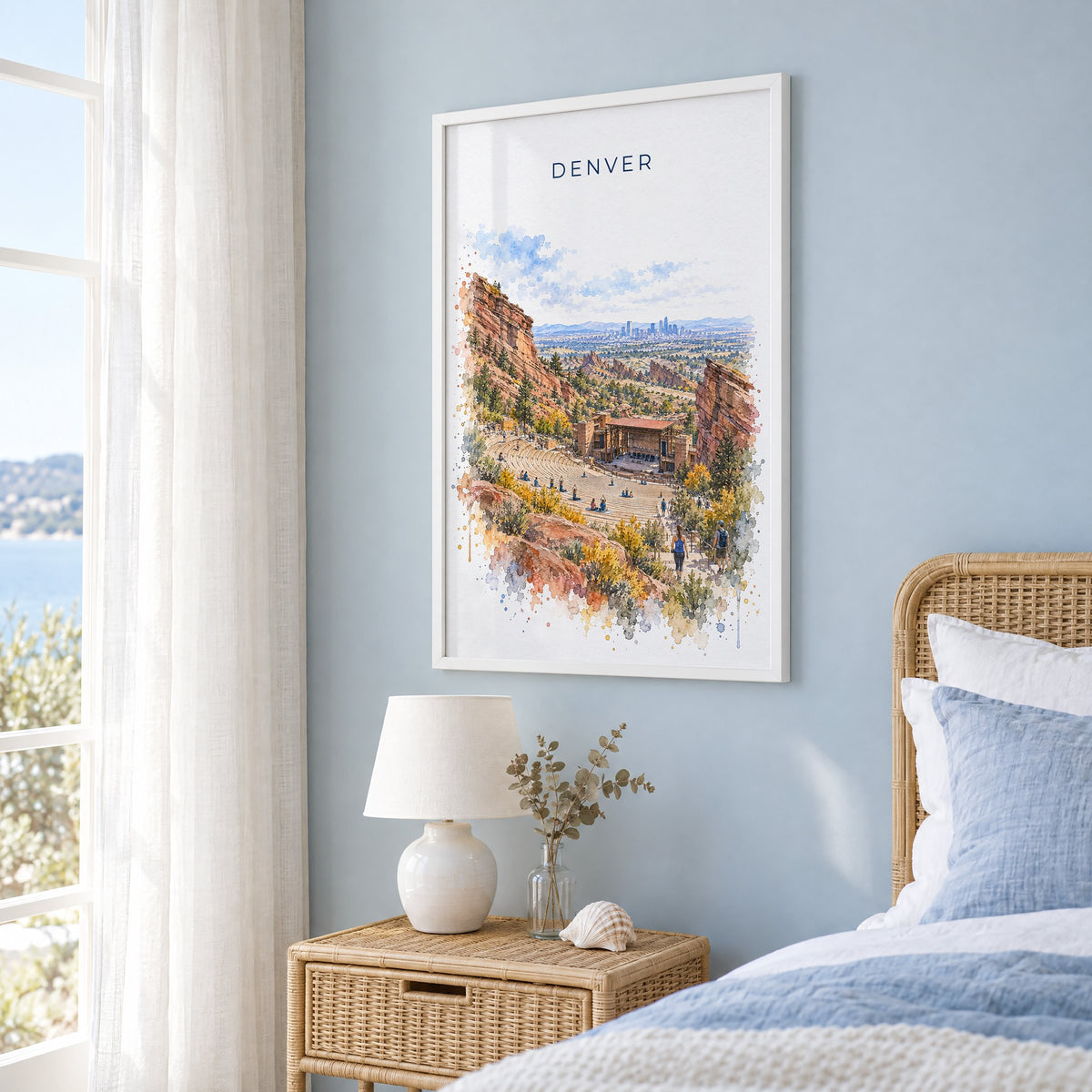 Red Rocks Amphitheatre watercolor print in a white frame on a pale blue wall beside a rattan bed in a coastal bedroom