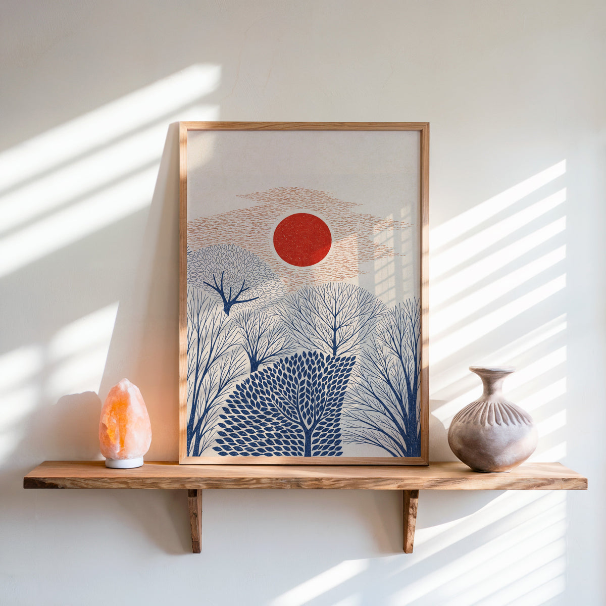 red sun over navy trees print in an oak frame on a live-edge wall shelf with a salt lamp and clay vase