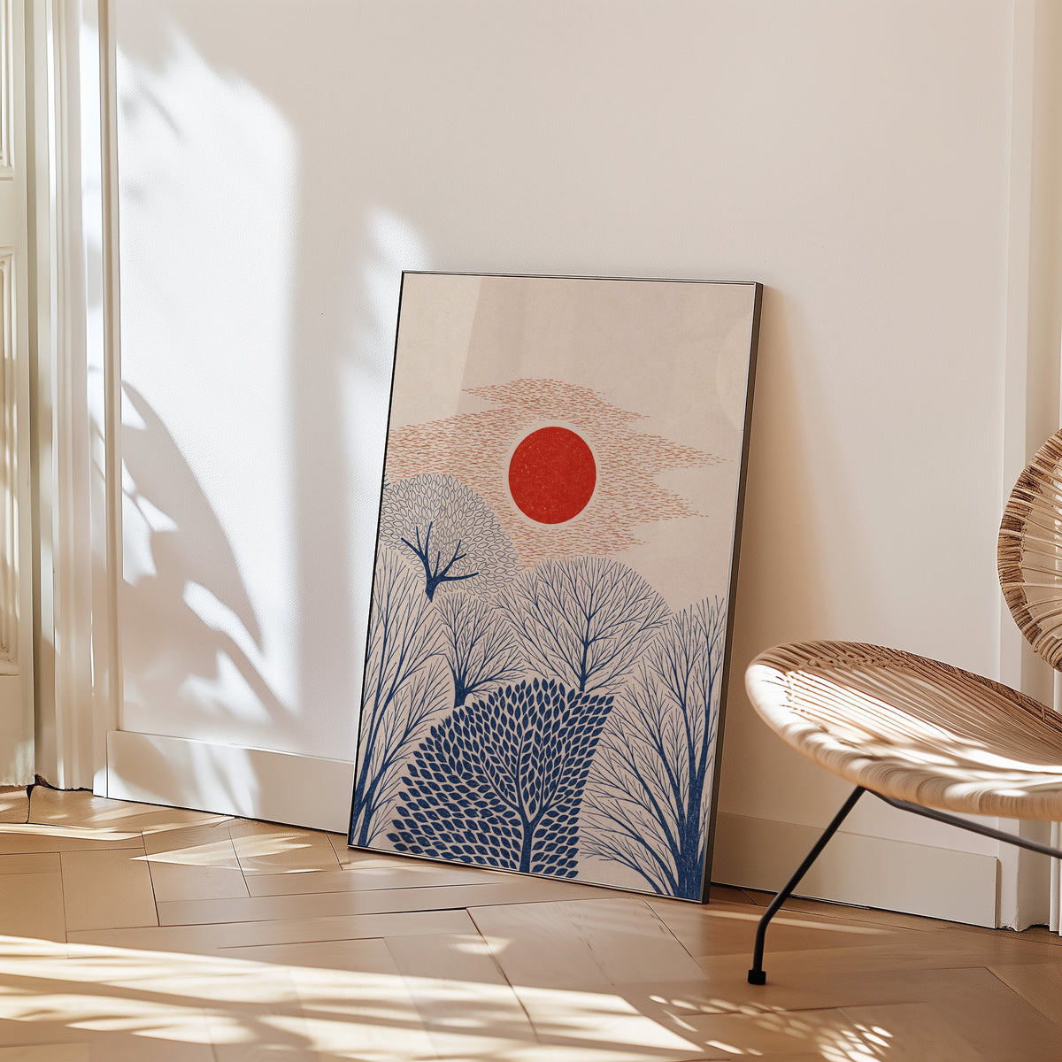 red sun over navy trees print in a black frame leaning on a herringbone floor beside a woven rattan chair