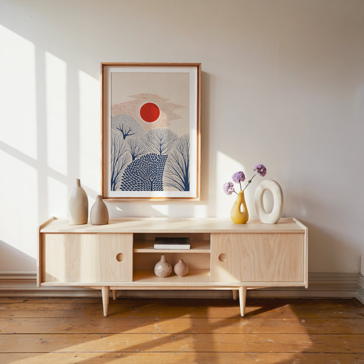 red sun over navy trees print shown in a wood frame with a white mat above a pale wood media console
