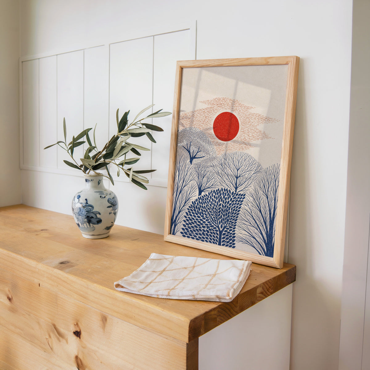 red sun over navy trees print in a pine frame on a wooden counter beside a blue and white porcelain vase