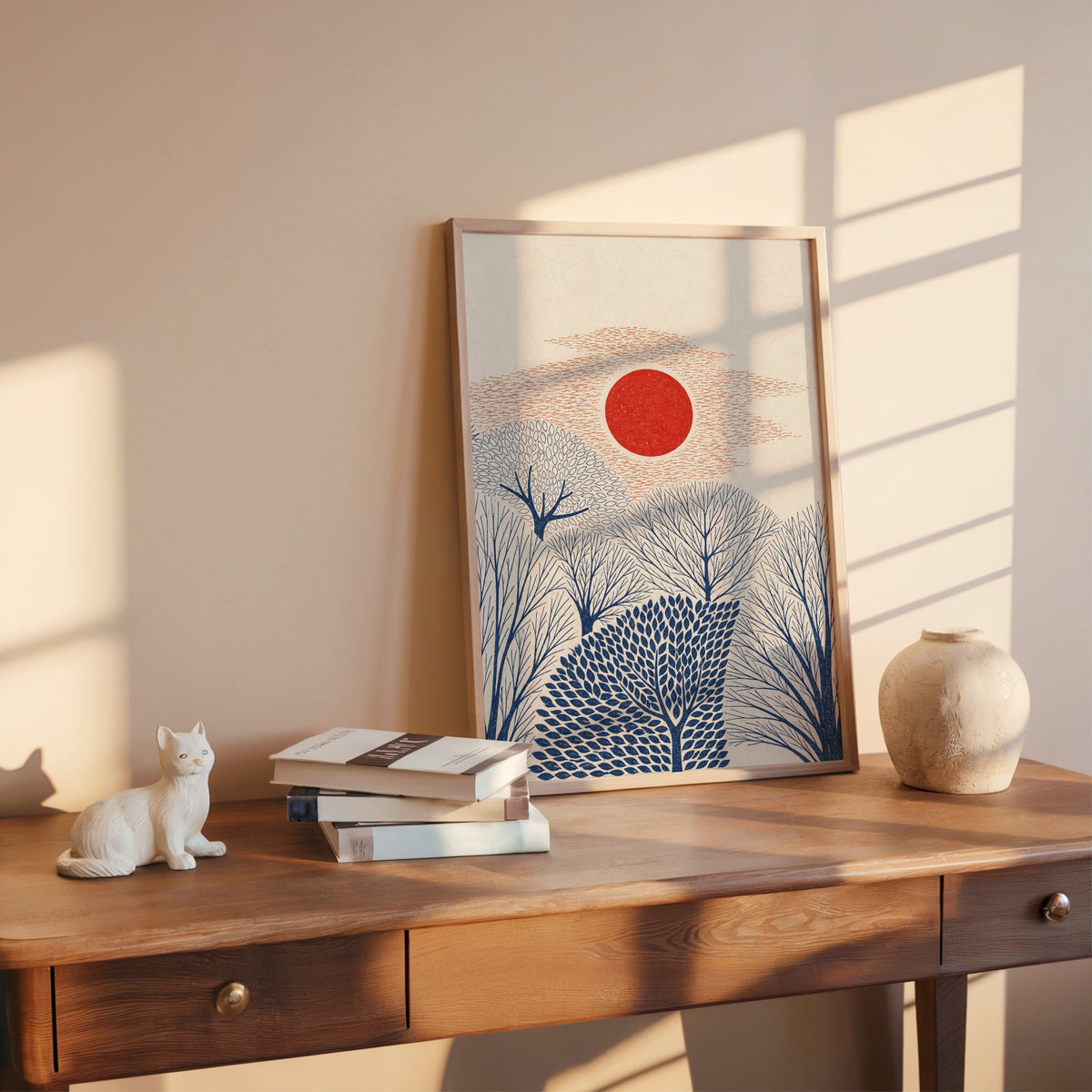 red sun over navy trees print shown in a rose gold frame leaning on a wooden writing desk with books