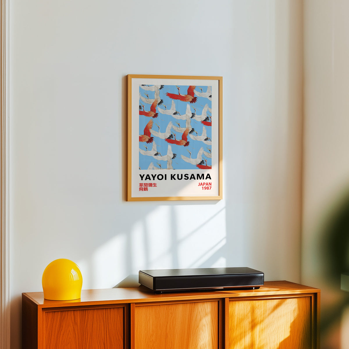Yayoi Kusama inspired red and white flying cranes print in an oak frame over a teak sideboard with a yellow lamp