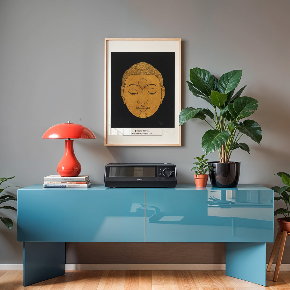 Buddha head print in an oak frame above a teal sideboard with a red lamp