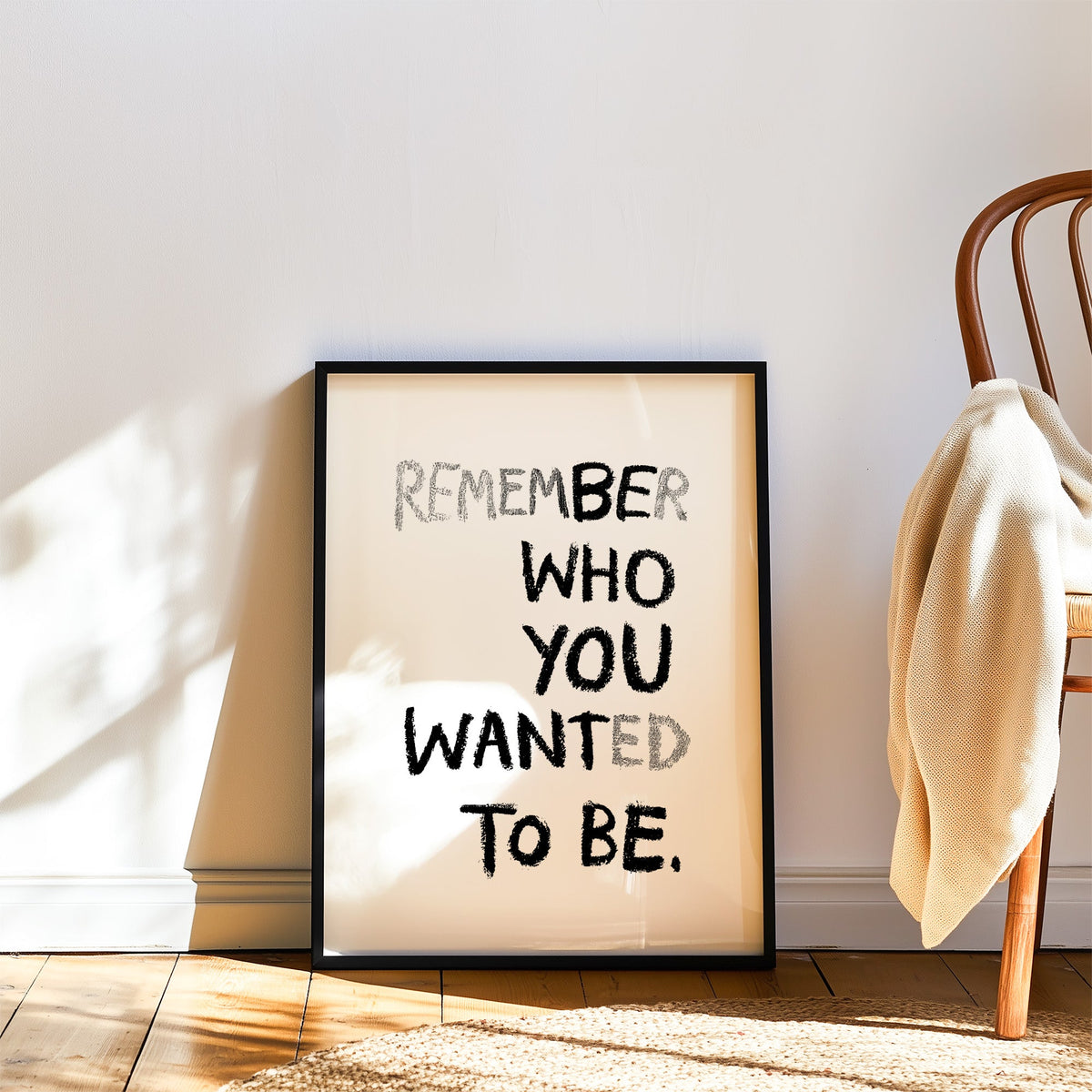 Crayon lettering remember who you wanted to be print in a black frame leaning on a wood floor beside a bentwood chair