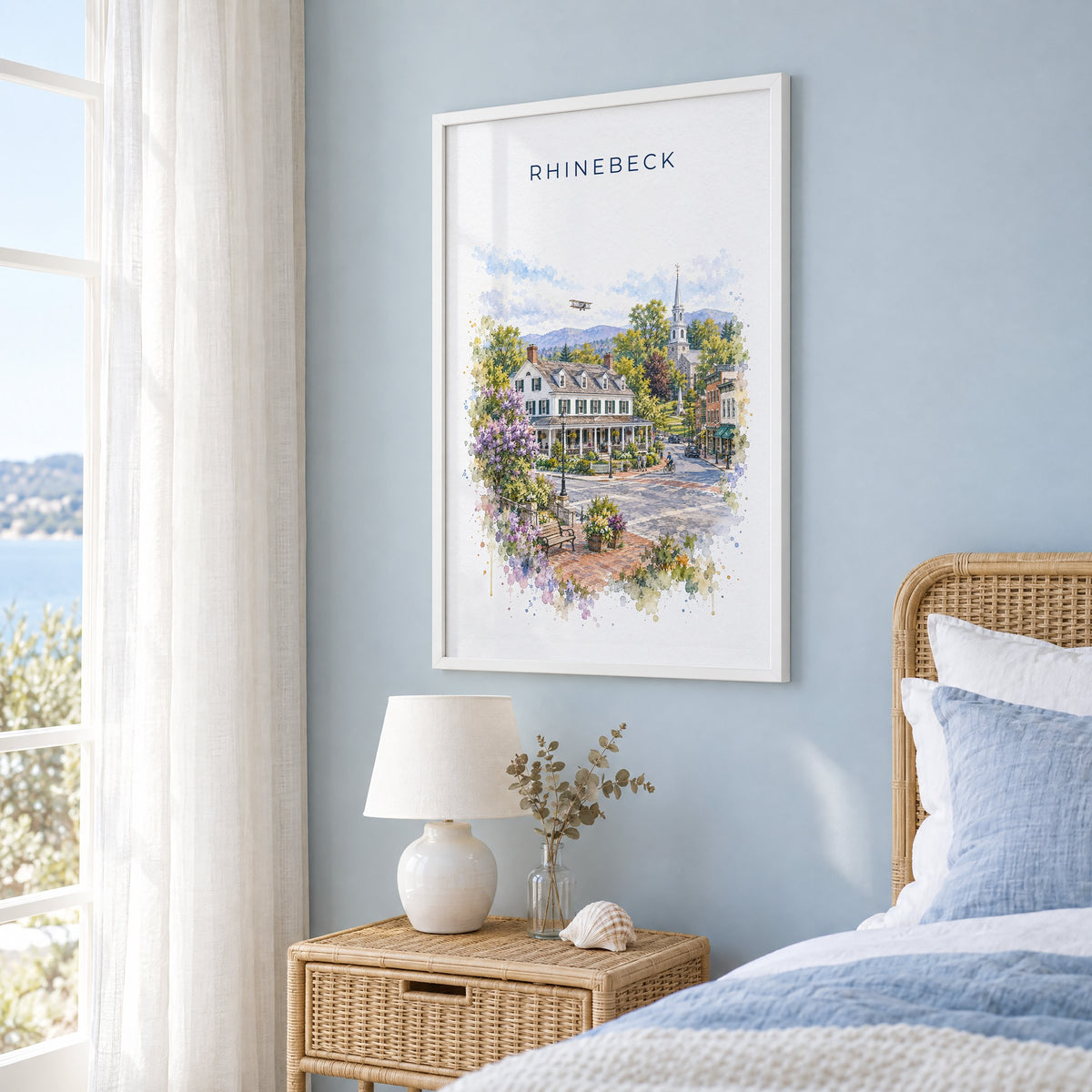 Rhinebeck, New York watercolor print in a white frame on a pale blue wall beside a rattan bed in a coastal bedroom