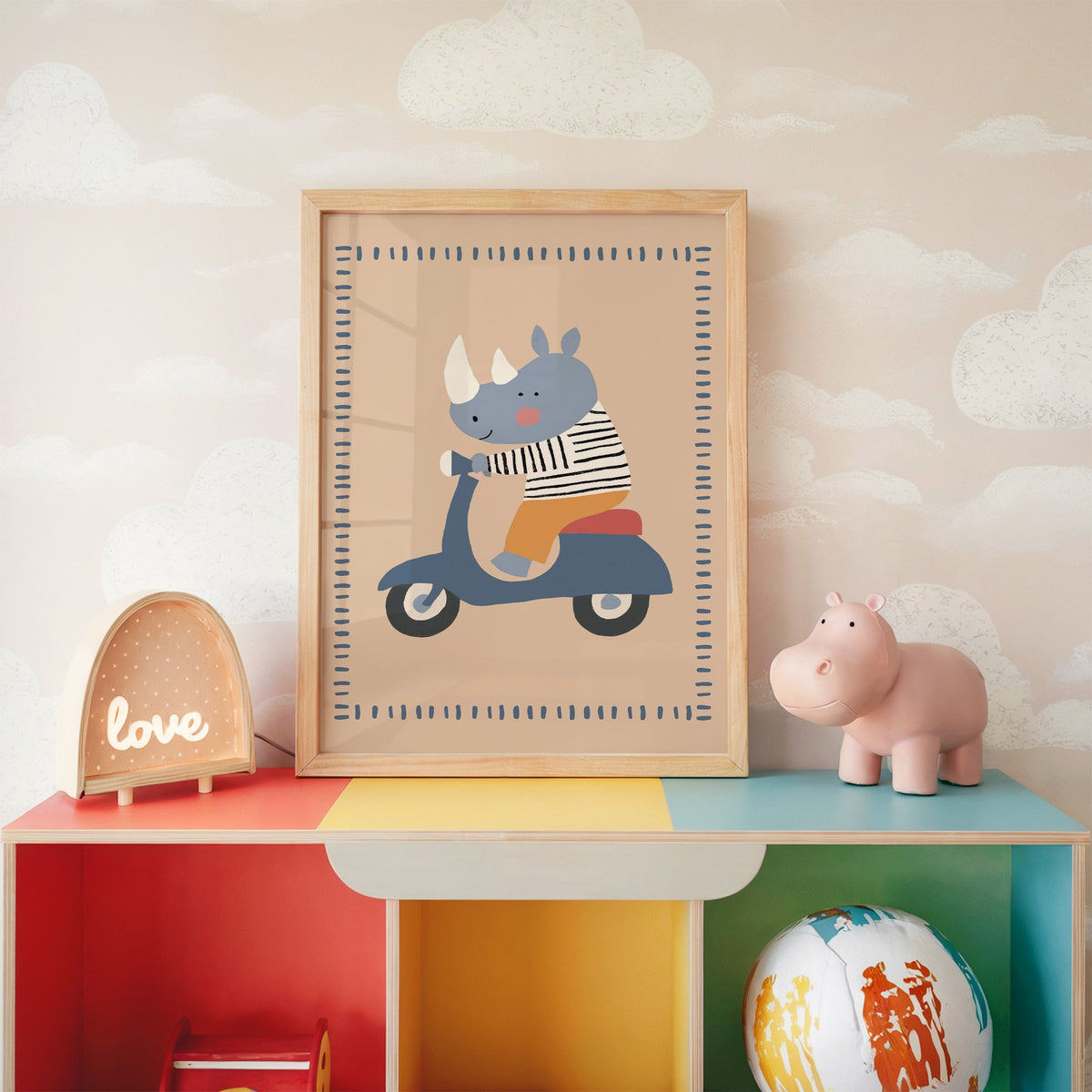 Rhino on a blue scooter nursery print in an oak frame on a rainbow toy shelf against cloud wallpaper