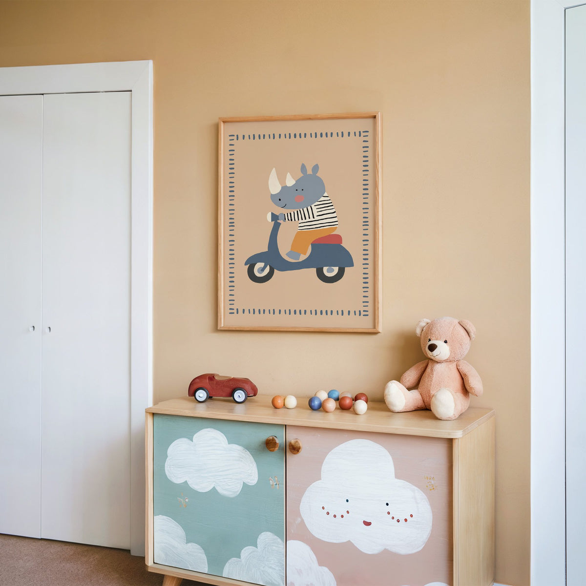 Rhino on a blue scooter nursery print in an oak frame on a peach wall above a cloud-painted cabinet