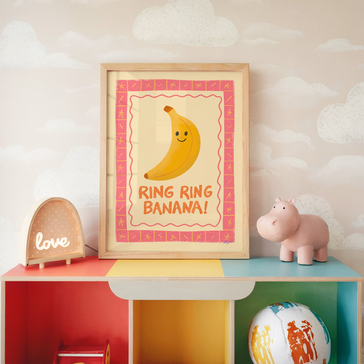 Smiling banana pink border nursery print in an oak frame on a rainbow toy shelf against cloud wallpaper
