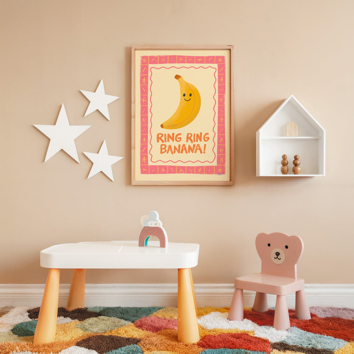 Smiling banana pink border nursery print in an oak frame on a peach wall above a kids table with wall stars