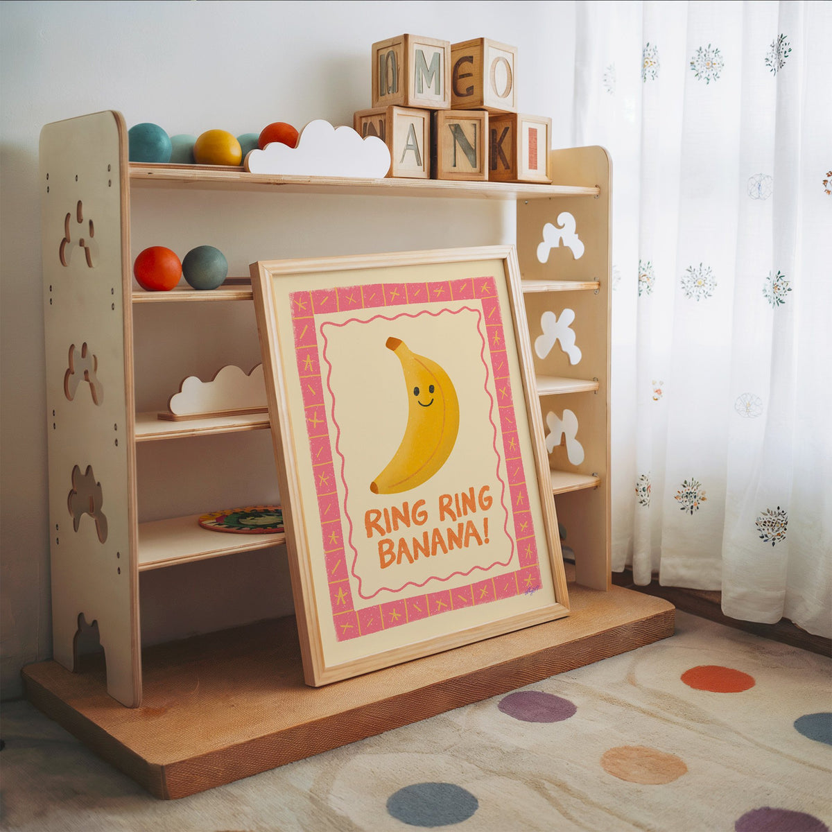 Smiling banana pink border nursery print in an oak frame leaning on a toy shelf with wooden letter blocks