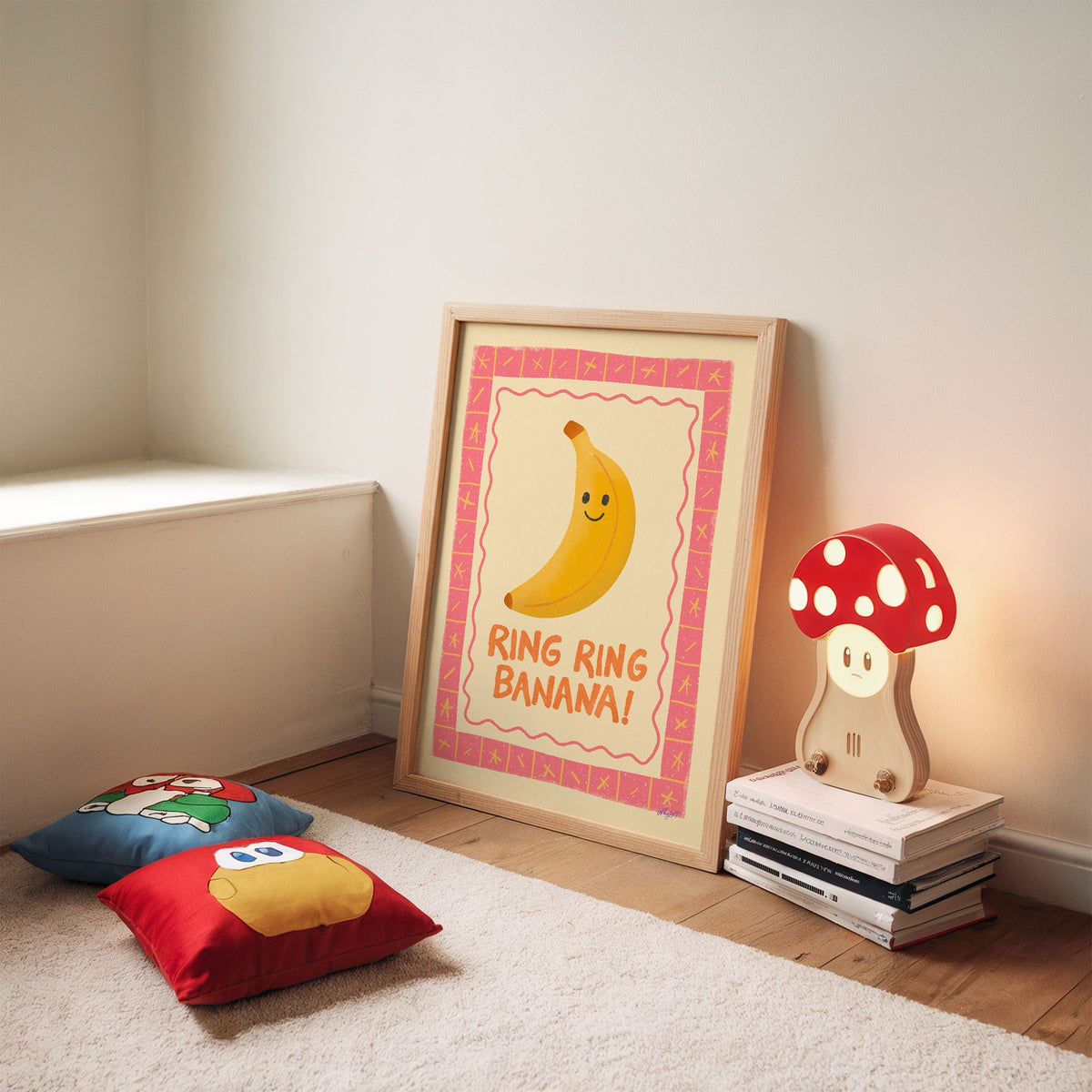 Smiling banana pink border nursery print in an oak frame leaning on a rug beside a mushroom lamp and floor cushions