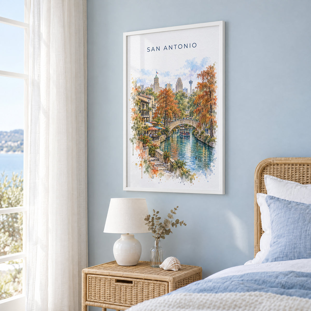 San Antonio River Walk watercolor print in a white frame on a pale blue wall beside a rattan bed in a coastal bedroom