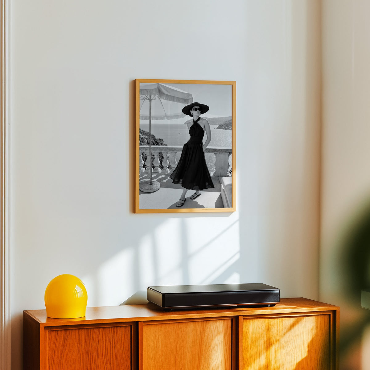 Black and white woman in halter dress on terrace print in an oak frame above a teak sideboard with a yellow glass lamp