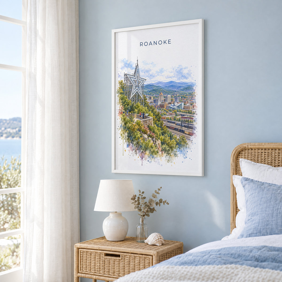 Roanoke watercolor print in a white frame on a pale blue wall beside a rattan bed in a coastal bedroom