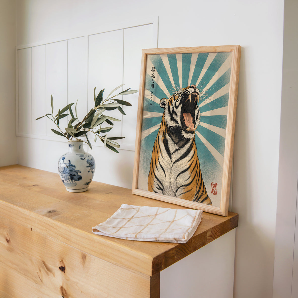 roaring tiger with teal sunburst print in a pine frame on a wooden counter beside a blue and white porcelain vase