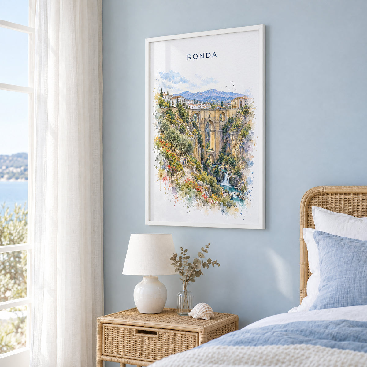 Ronda watercolor print in a white frame on a pale blue wall beside a rattan bed in a coastal bedroom