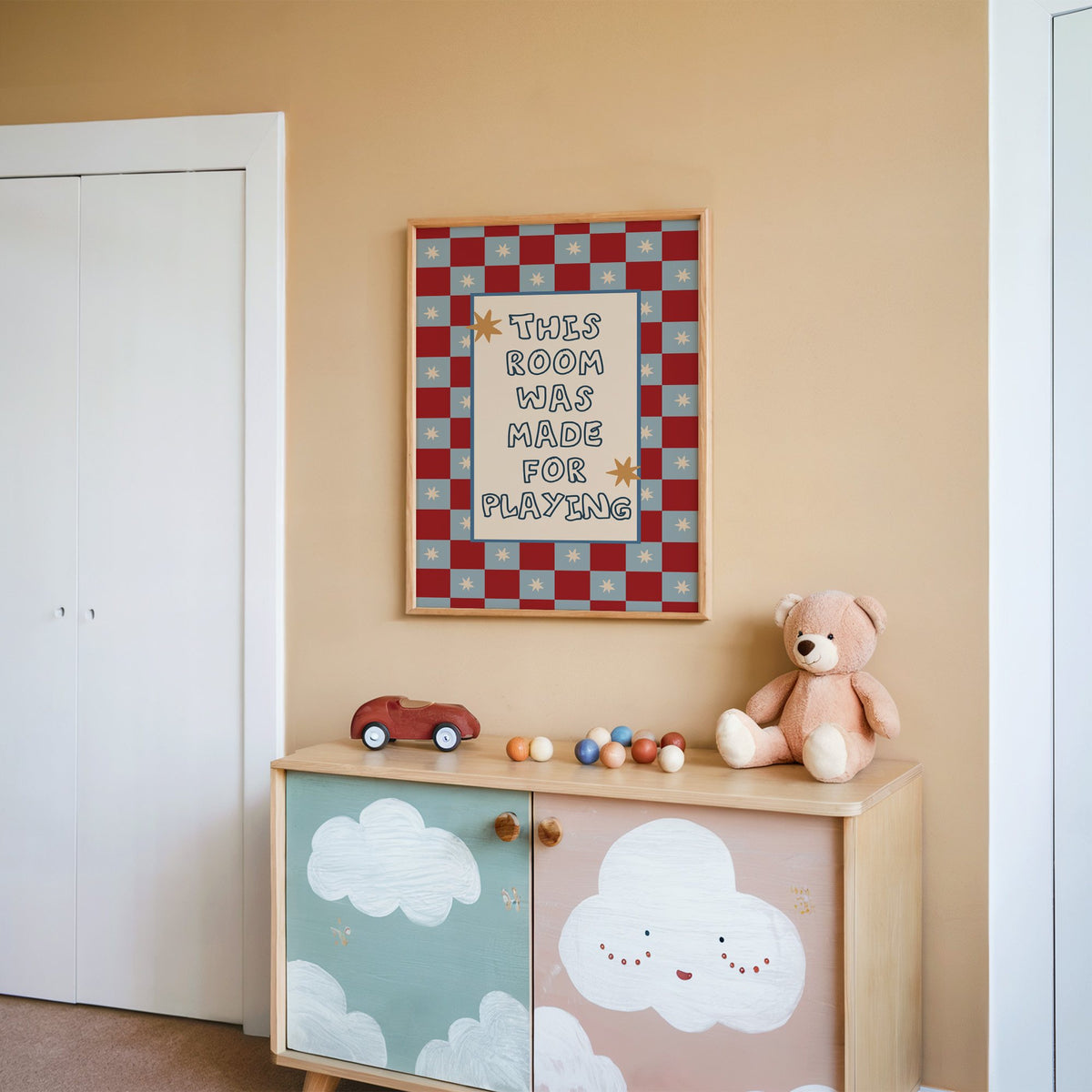 Room made for playing checkered nursery print in an oak frame on a peach wall above a cloud-painted cabinet