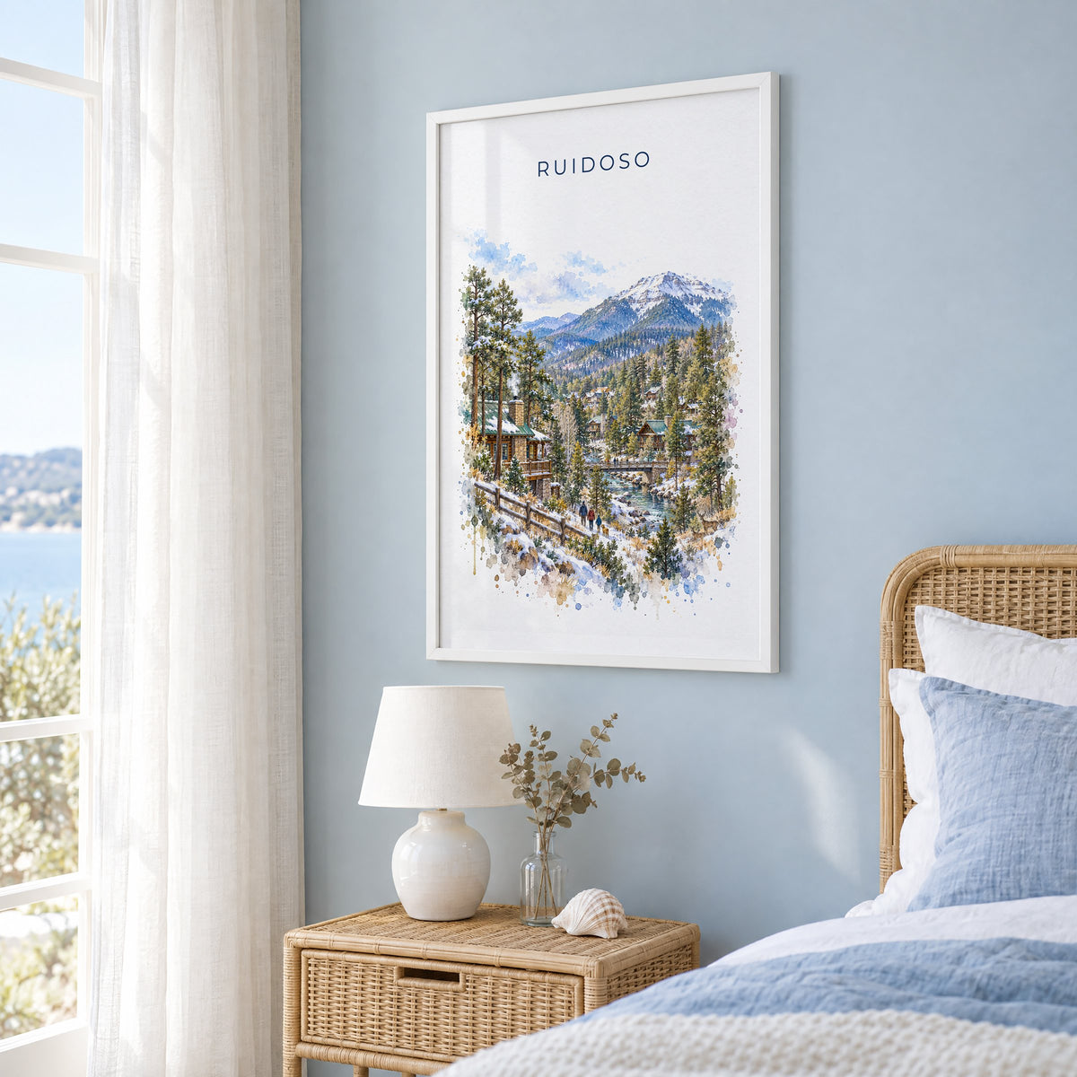 Ruidoso, New Mexico watercolor print in a white frame on a pale blue wall beside a rattan bed in a coastal bedroom