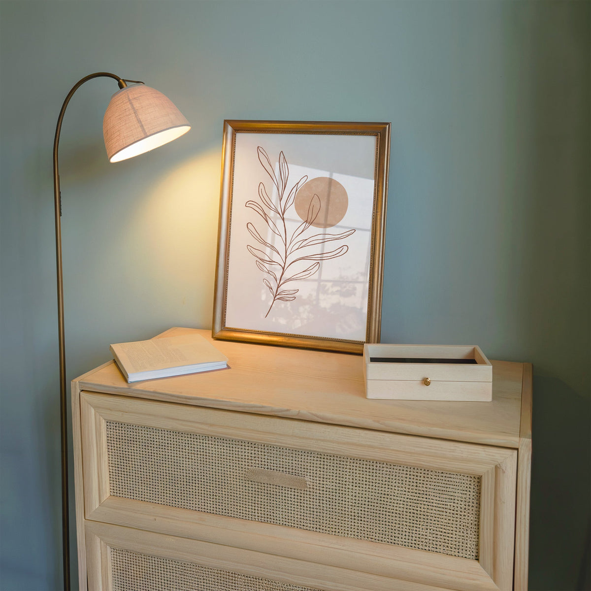 Rust line leaf branch and peach sun botanical print shown in a gold frame on a cane dresser lit by a floor lamp