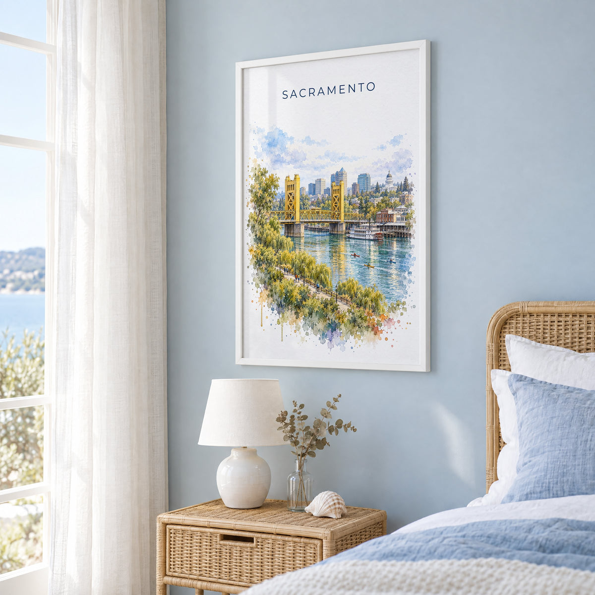 Sacramento watercolor print in a white frame on a pale blue wall beside a rattan bed in a coastal bedroom