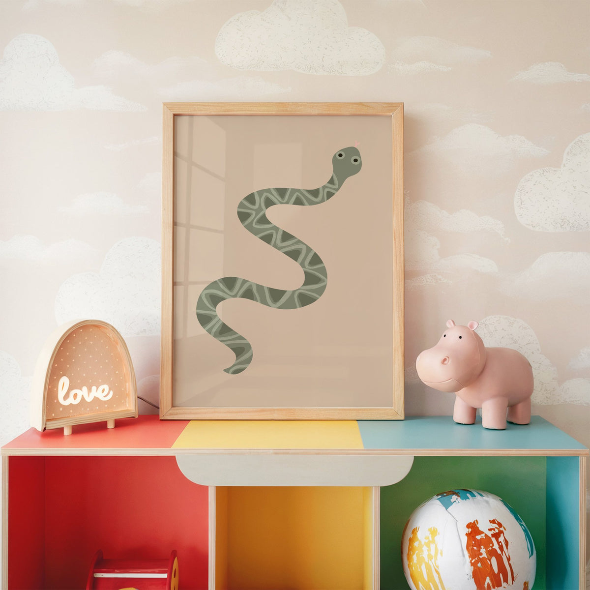 Sage green snake nursery print in an oak frame on a rainbow toy shelf against cloud wallpaper