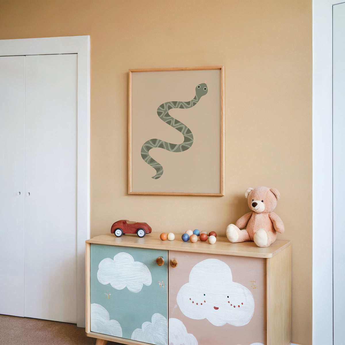 Sage green snake nursery print in an oak frame on a peach wall above a cloud-painted cabinet