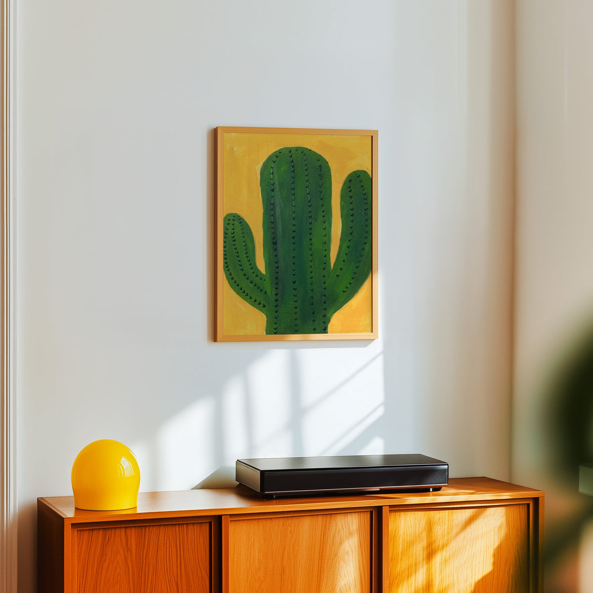 Mustard and green cactus print in a light oak frame above a teak sideboard with a yellow lamp