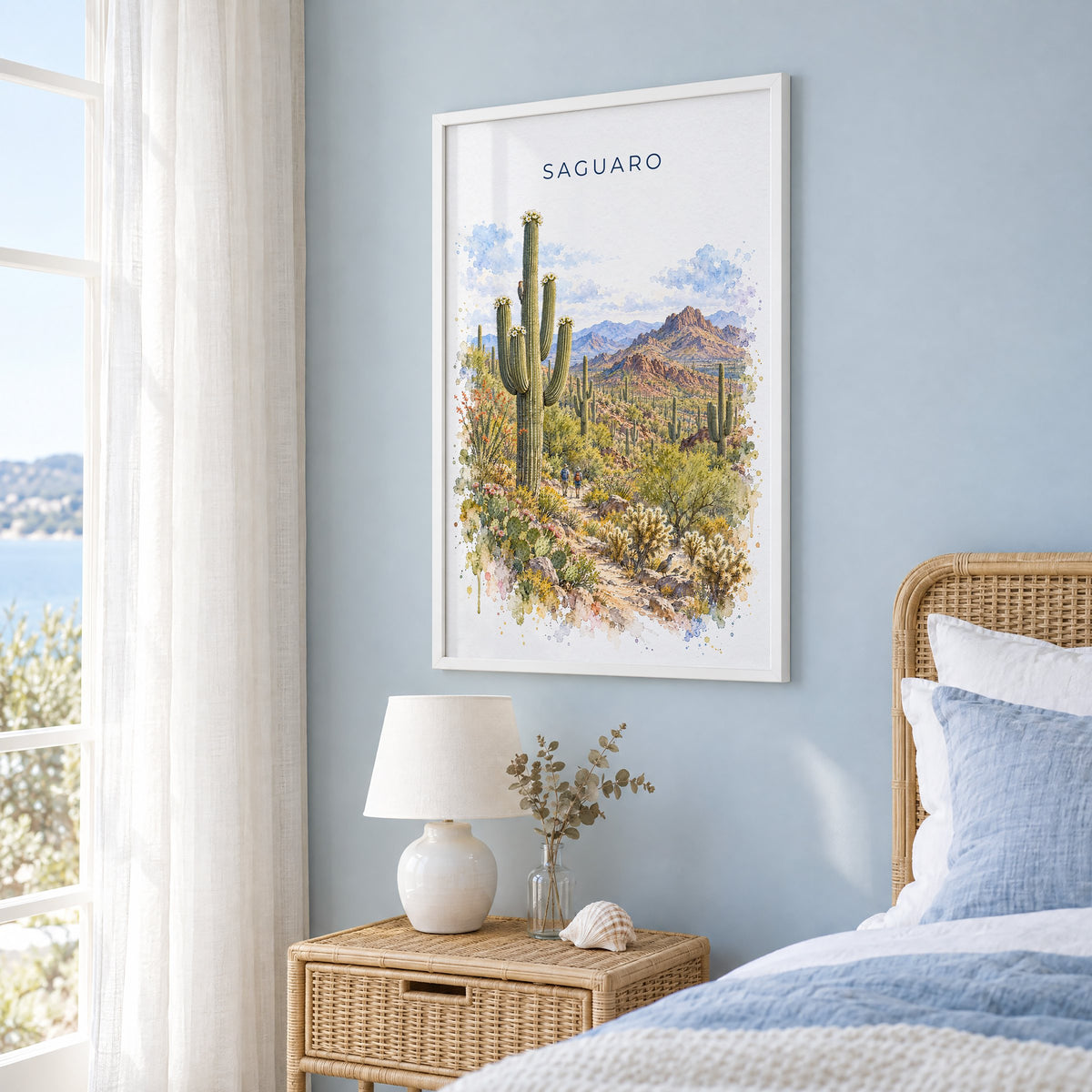 Saguaro National Park watercolor print in a white frame on a pale blue wall beside a rattan bed in a coastal bedroom